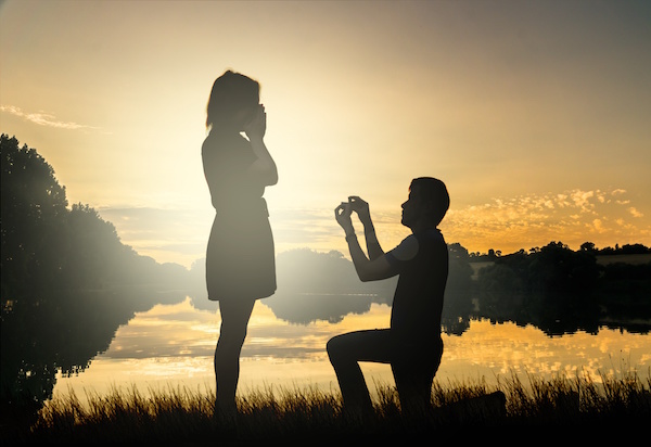 Wedding proposal concept. Young couple have dating at sun set. Man in love is kneeling on knees and giving wedding ring to surprised woman.